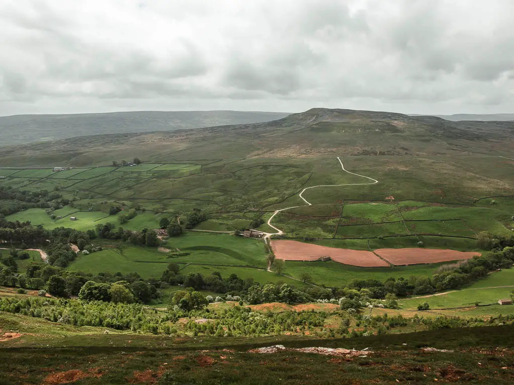 Looking down into the valley on the Fremington Edge walk. The valley is filled with green fields, with a hill and peak rising up on the other side, with a road curving up it.