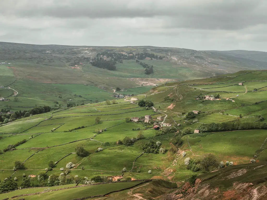 Looking down and across the green valley fields, with a small settlement of houses at the bottom, on the walk along Fremington Edge.