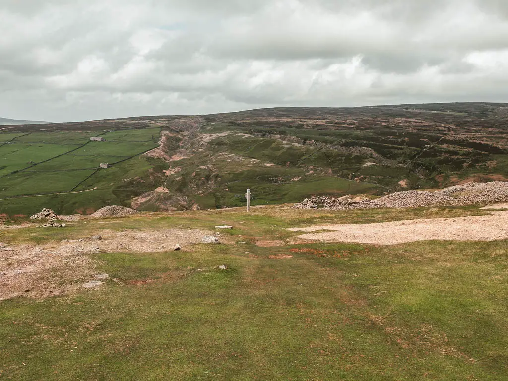A grassy hilltop with patches of gravel, and a trail signpost ahead, and a hill rising up in the distance.