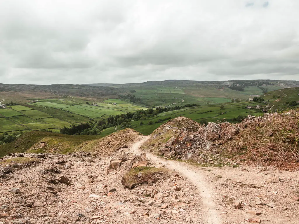 A path curving ahead through the rocky ground, with a view to the green undulating hills ahead in the distance.