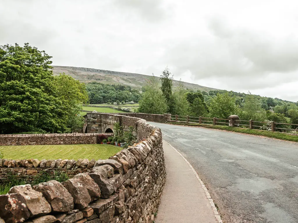 A road leading over a stone walled bridge, with Fremington edge visible ahead in the distance. There are lots of green leafy trees on the other side of the bridge.