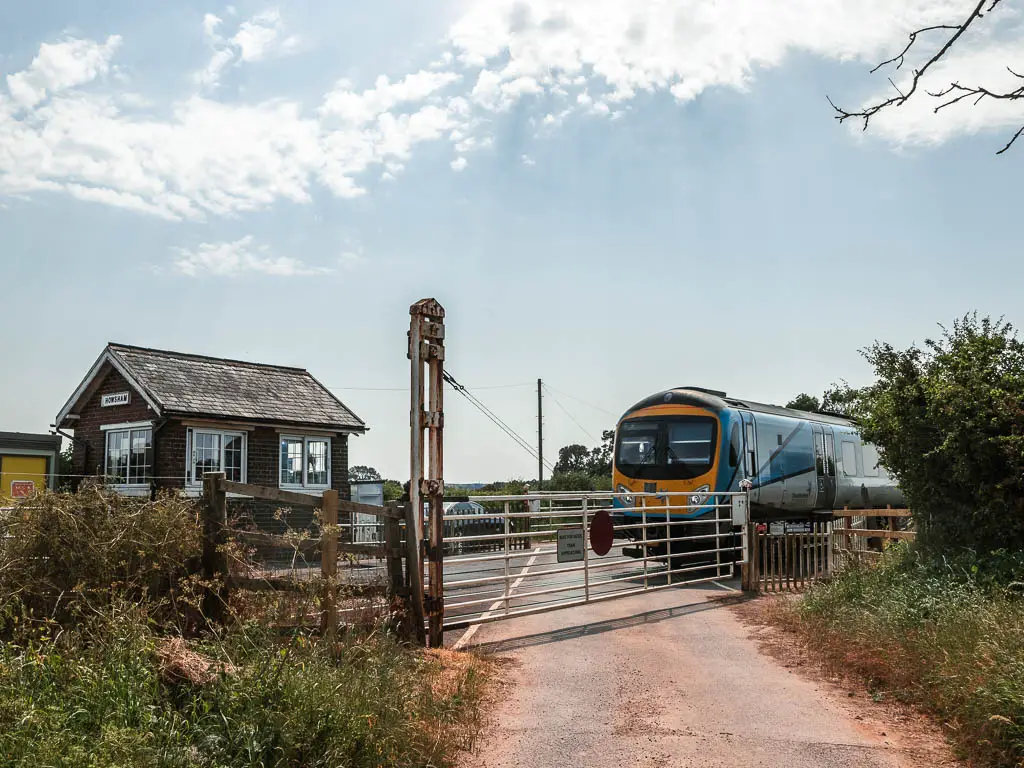 The road leading to a closed white gate next to the railway line, part way through the Kirkham Priory circular walk. There is a train on the track, and a small brick walled train building on the other side of the tracks.