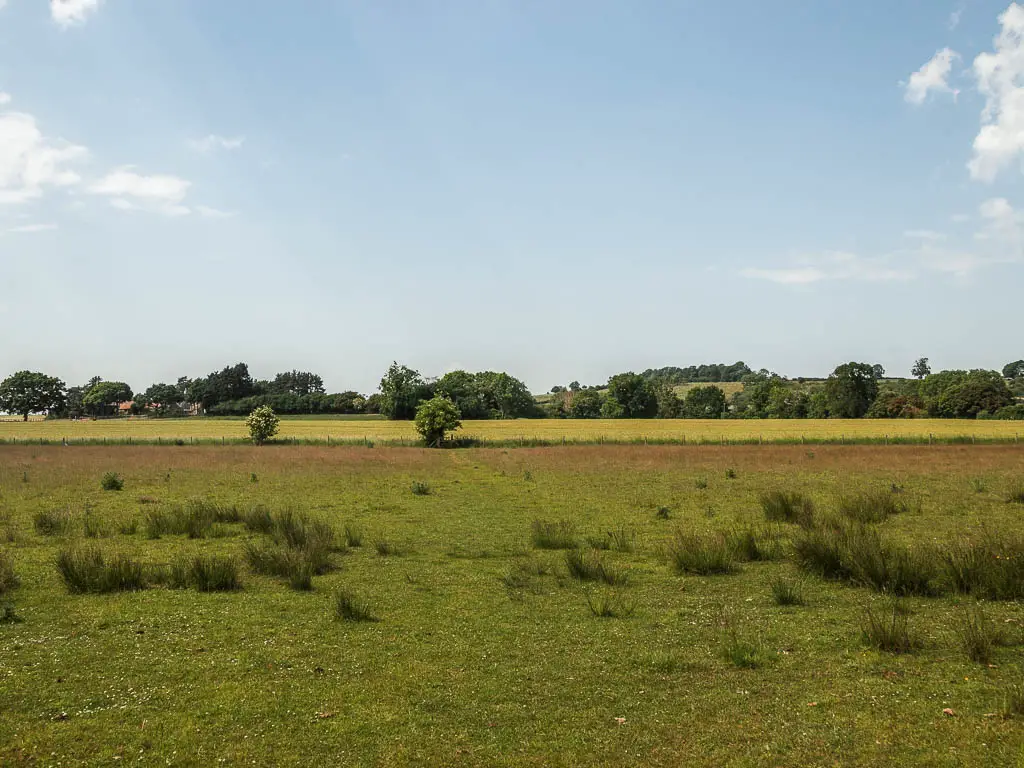 A large green grass field, leading to a lighter green field, part way through the Kirkham Priory circular walk. There are tufts of grass in the field. 