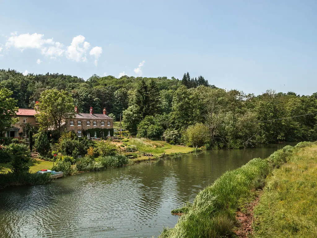 Looking across the river, with cottages on the other side, with a mass of woodland behind them, at the start of the walk from Kirkham Priory.