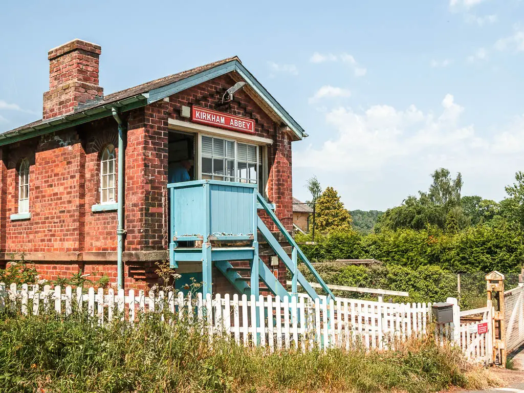 A small brick walled station building, with light blue steps leading up to the door. There is a red sign over the4 door saying 'Kirkham Abbey' in white writing.