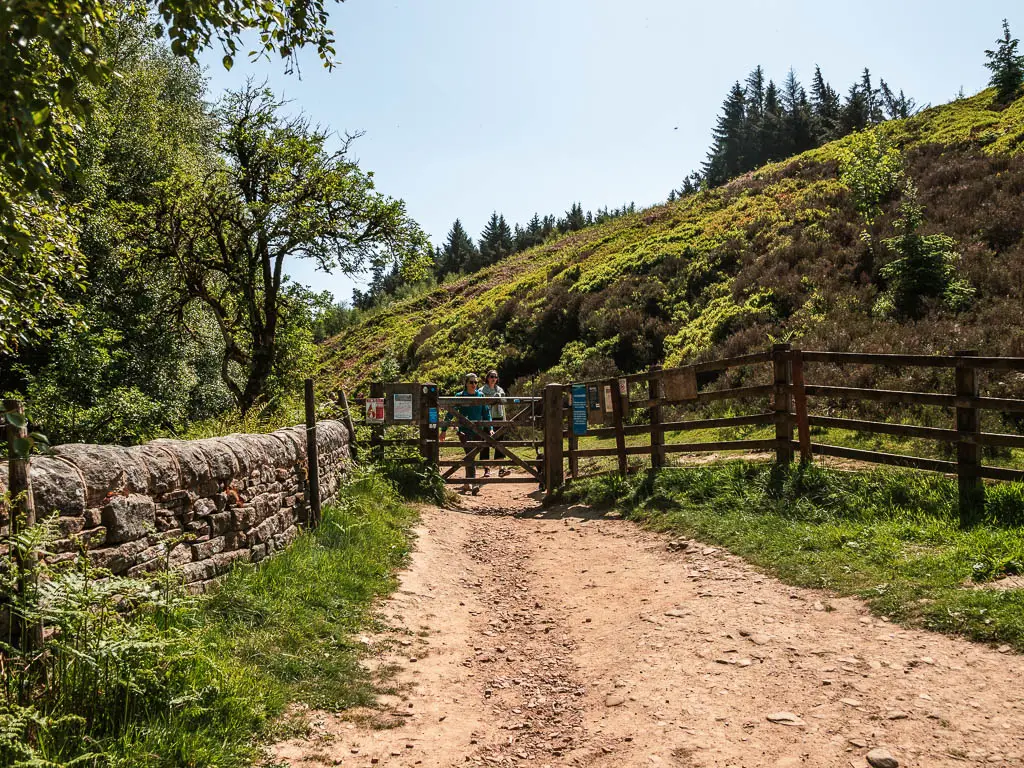 A stoney rugged path leading to a wooden gate. There is a stone wall on the left, and a moorland hill ahead on the other side of the gate and fence.