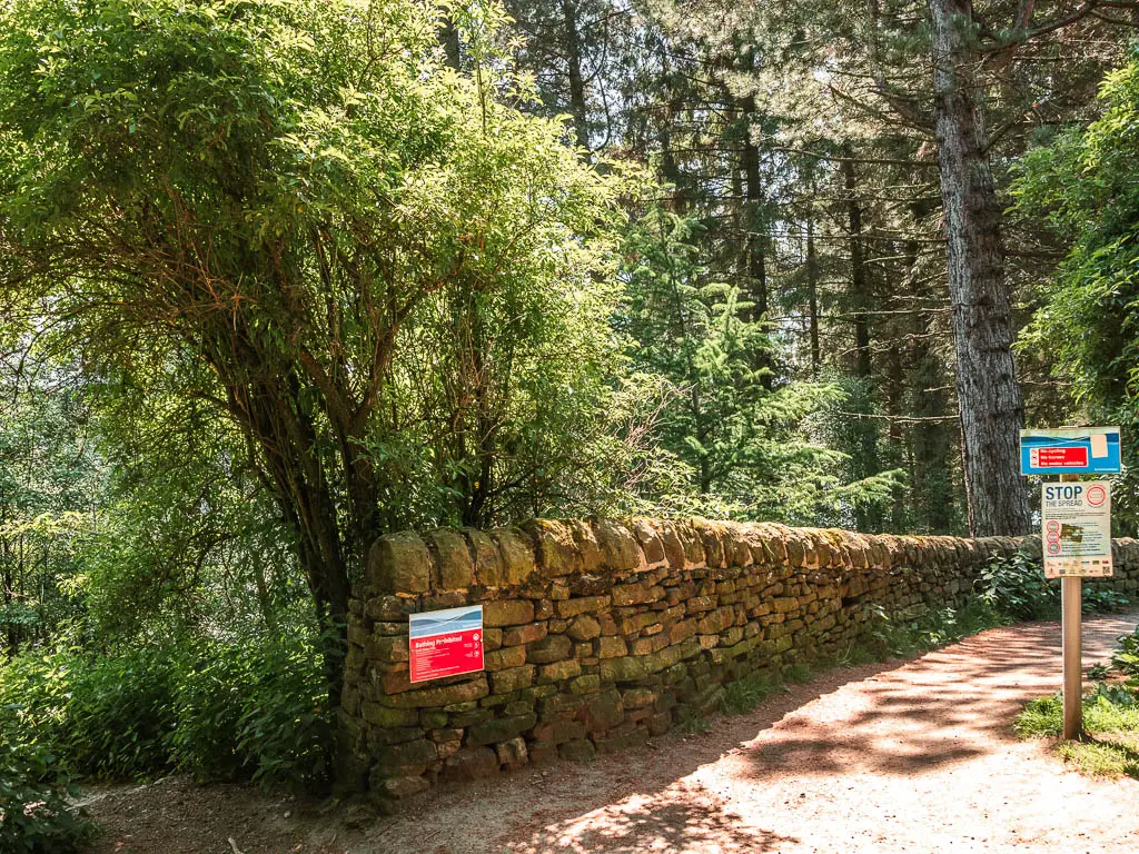 A path leading down to the right, with a stone wall running along the left side. There are green leafy trees on the other side of the wall.