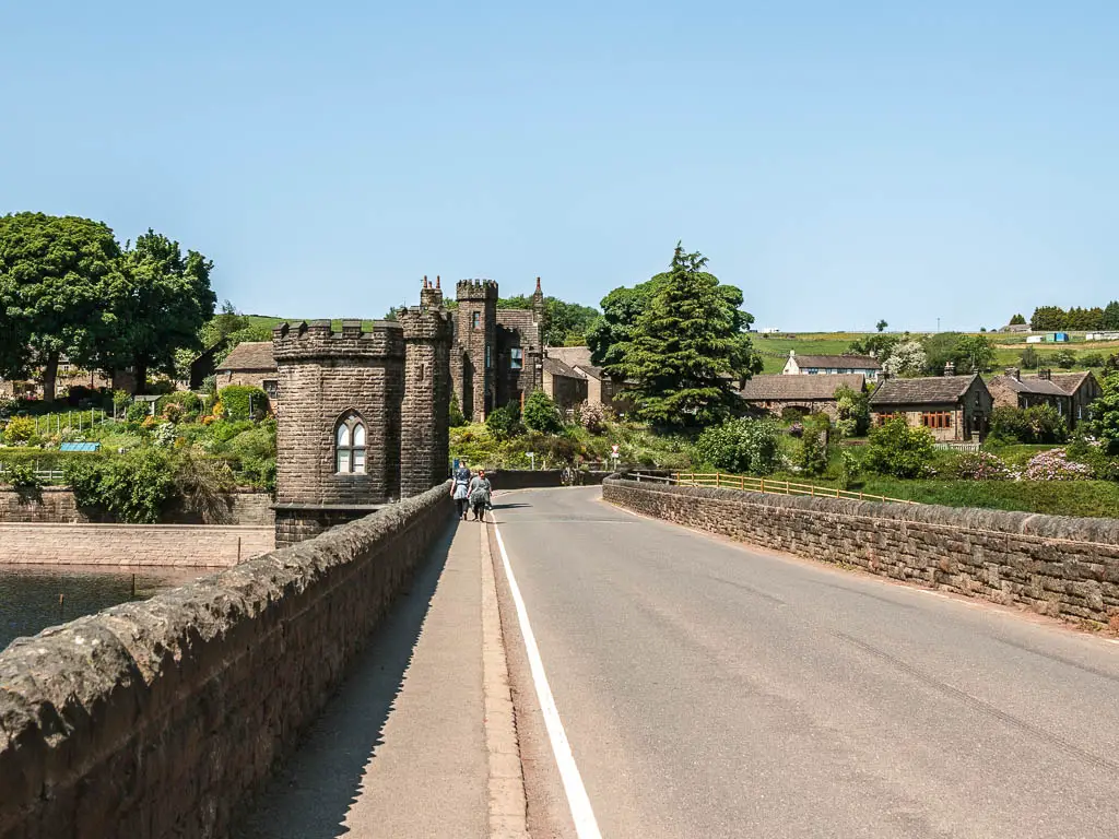 A road bridge, with a fort type structure ahead on the left. There are cute cottages and trees on the other end of the road.