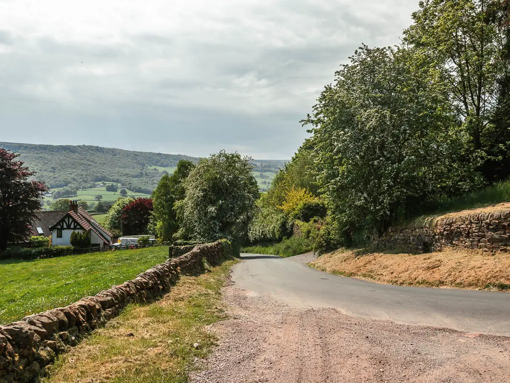 The gravel path leading onto the road which is lined with stone walls. there is a grass field on the other side of the left stone wall, and some rooftops visible on the other side of the field. further ahead, the road is lined with trees.