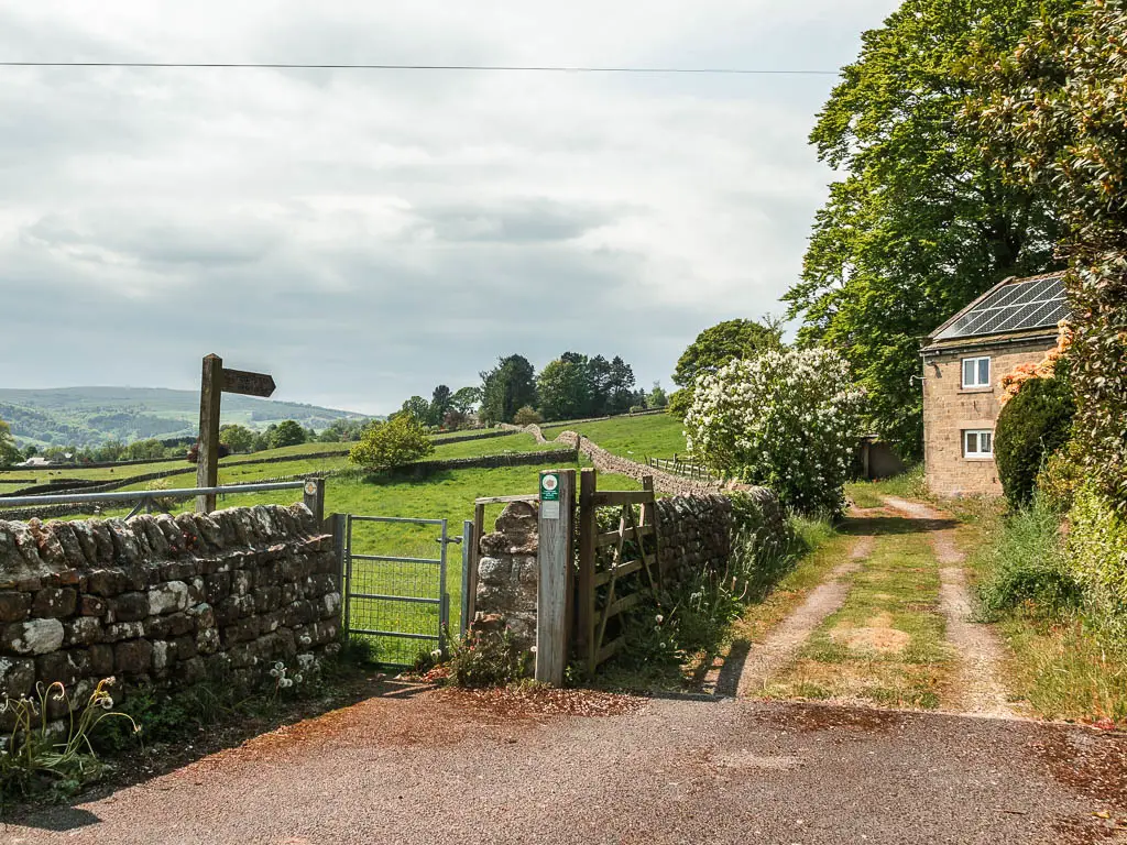 A metal gate pin the stone wall on the left, with a wooden trail sigpost pointing through it. There is a driveway leading to a house on the right.