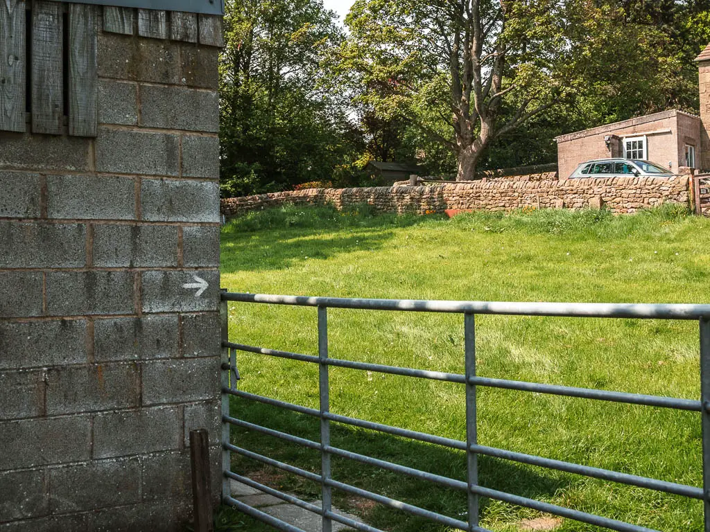 A metal gate with a shed on the left side. There is a white arrow on the shed wall, pointing through the gate into the small field.