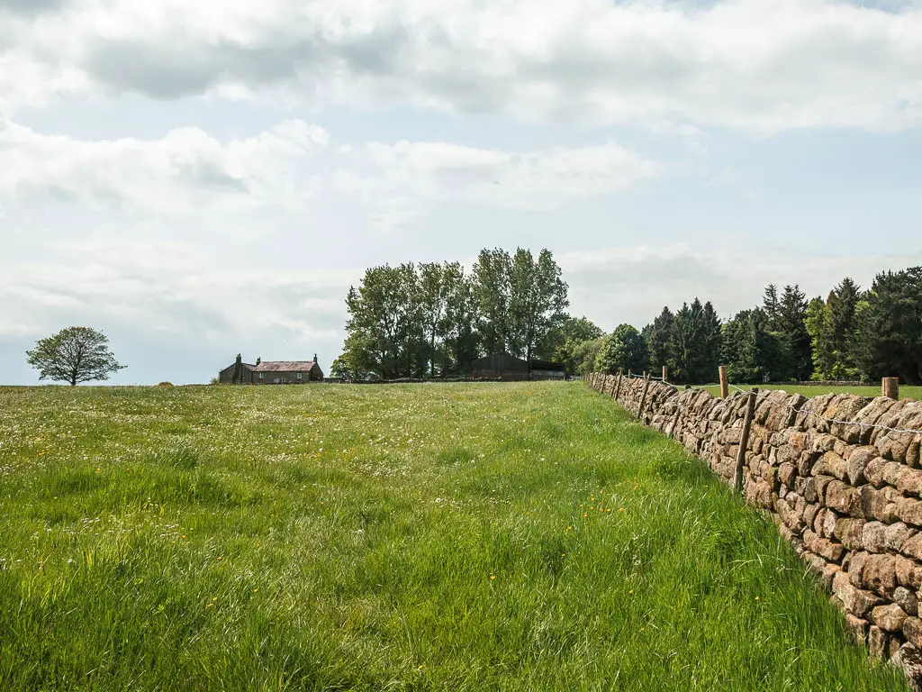 Looking up the grass field, with a stone all on the right and the rooftop of a farmhouse visible ahead to the left.