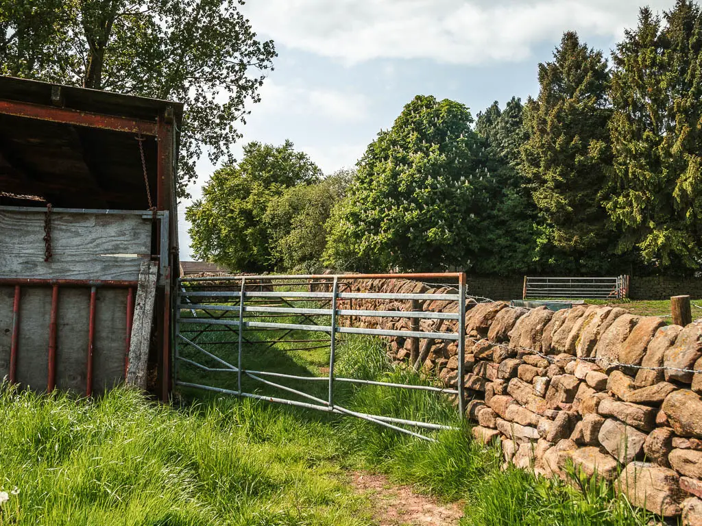 A partially broken metal gate with a farm building to the left and stone wall to the right.