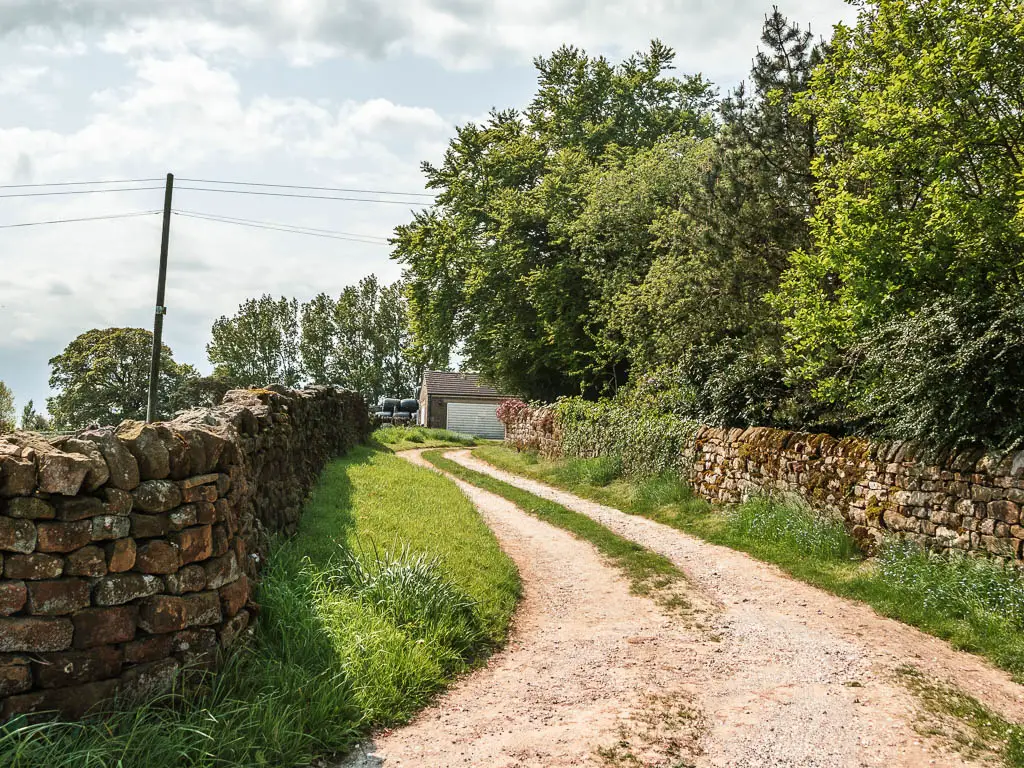 A driveway track, curving ahead, lined with stone walls. There are trees on the other side of the stone wall on the right, and shed visible at the end of the trail ahead.