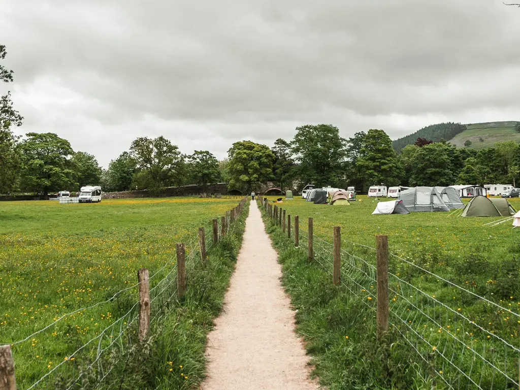 A long straight path, running between two large green fields. The field on the right is filled with tents. 