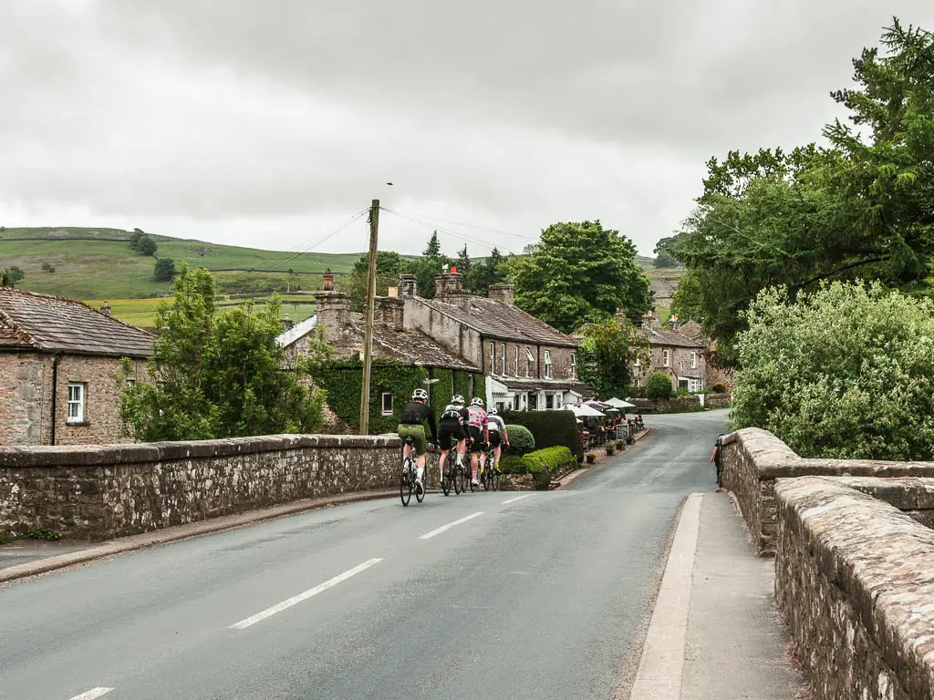 A bridge road with cyclists heading over it, part way through the Reeth circular walk. there are houses on the other side of the bridge.