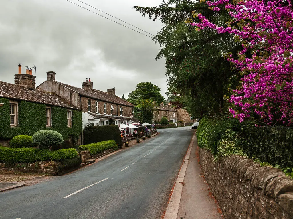 A road with a pavement and stone wall on the right, and trees with purple flowers leading over the wall, partway through the walk from Reeth. There is house covered in green ivy, on the other side of the road, and the pub is next to it. 