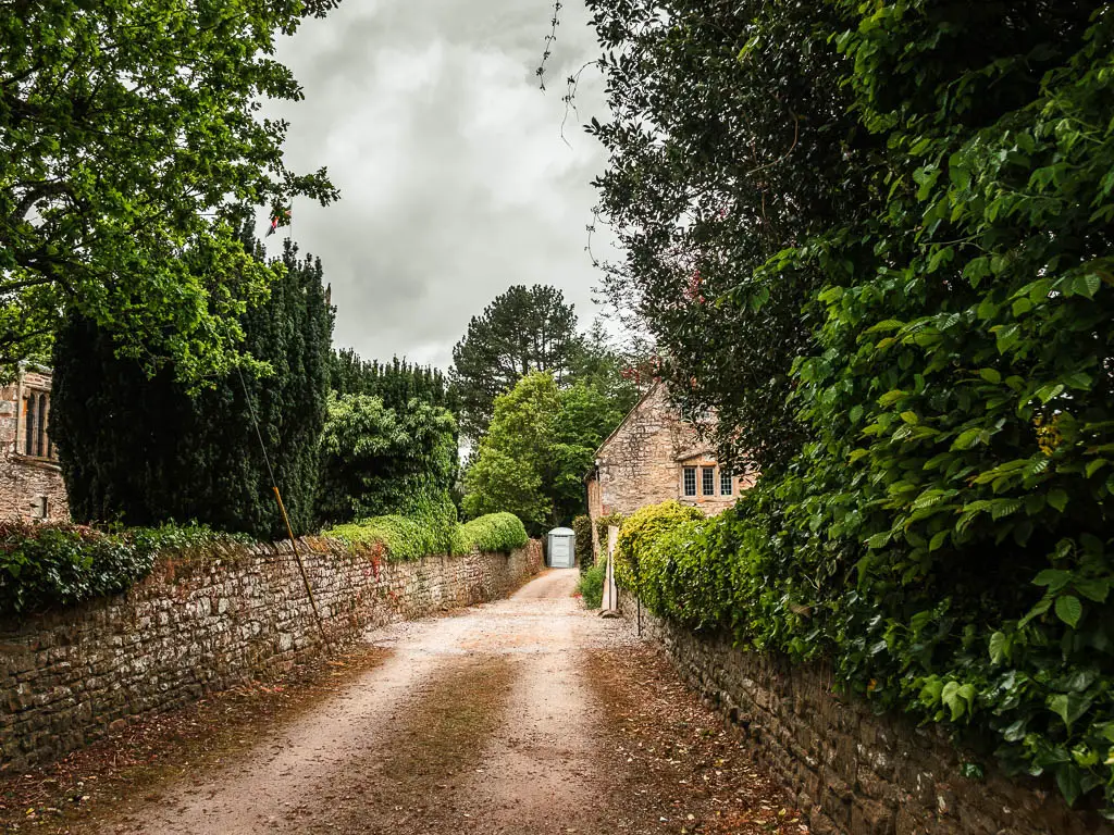 A wide driveway type road, lined with stone walls. there is a stone walled cottage ahead, with a porta-loo next to it. There are green leafed bushes and trees overhanging the right wall.