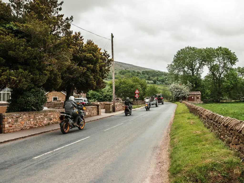 A road leading straight ahead, with tractors and motorbikes driving along it. There are houses on the left side of the road, and a grass bank and stone wall on the right.