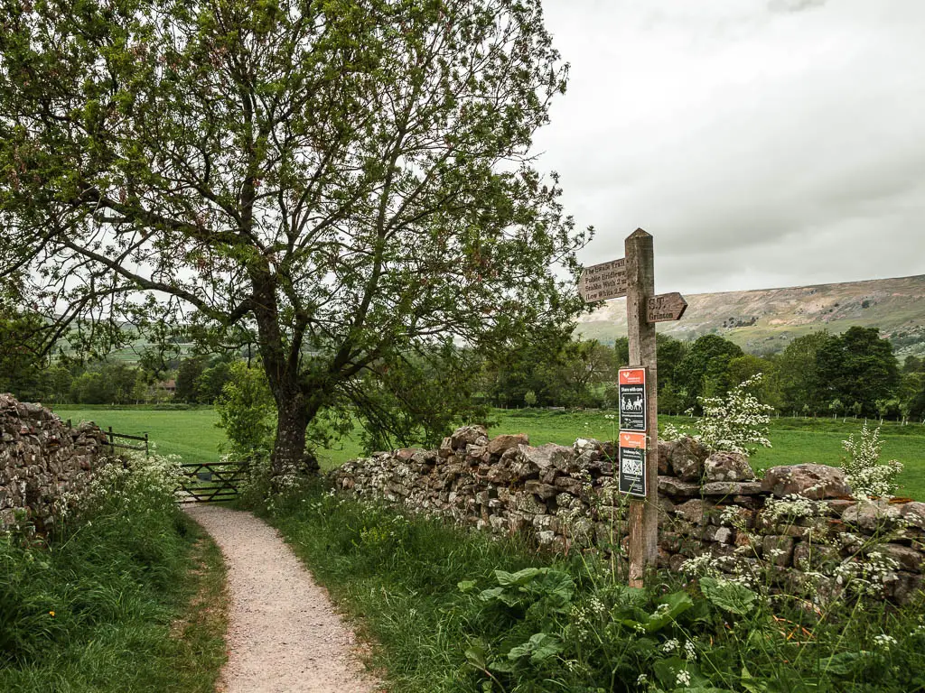 A wooden trail signpost pointing ahead along a gravel trail. There is a wooden gate on the end of the trail ahead. There is a stone wall to the right.