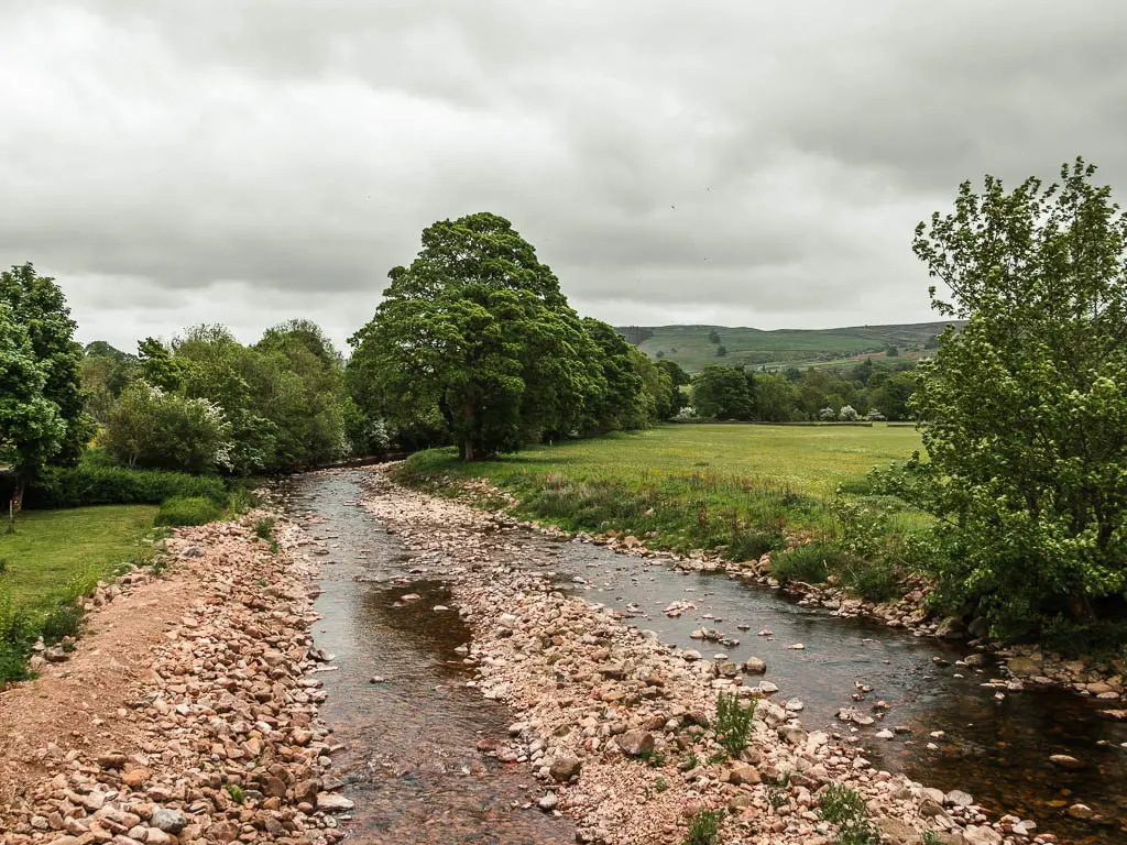 Looking along the river, filled with large stones and rocks, at the start of the walk out of Reeth. the river is lined with grass fields and a trees.