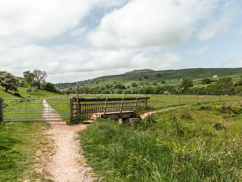 A gravel path on the left, leading to a small wooden bridge, on the walk to the suspension bridge near Reeth.