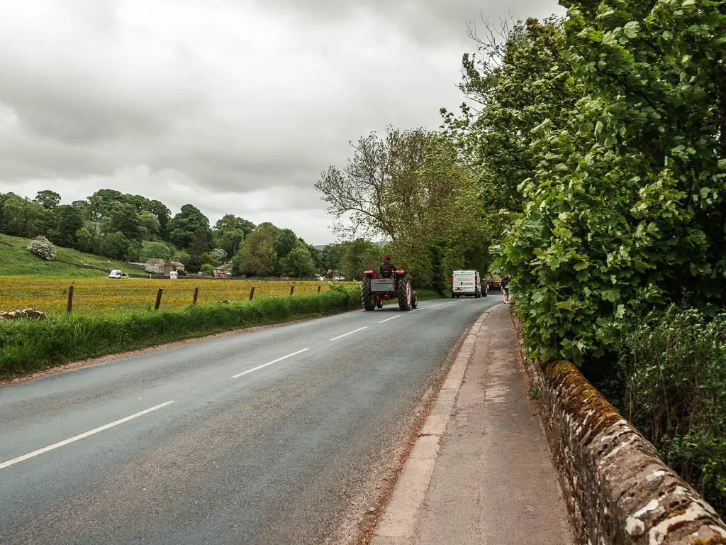 A road leading straight ahead, with a tractor driving on it. there is a pavement and stone wall on the right, and a field on the left.