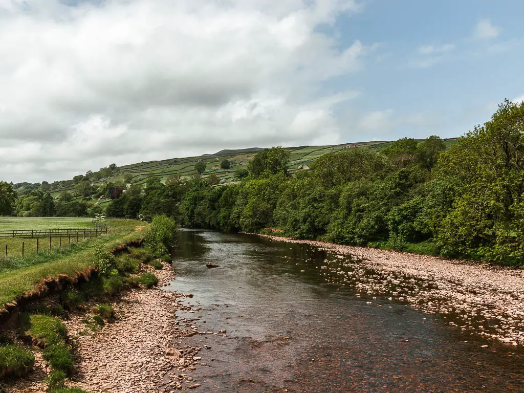 Looking along the river, when standing on there suspension bridge, on the walk back to Reeth. the river is lined with rocks, and a green grass field on the left, and masses of green leafy trees on the right.