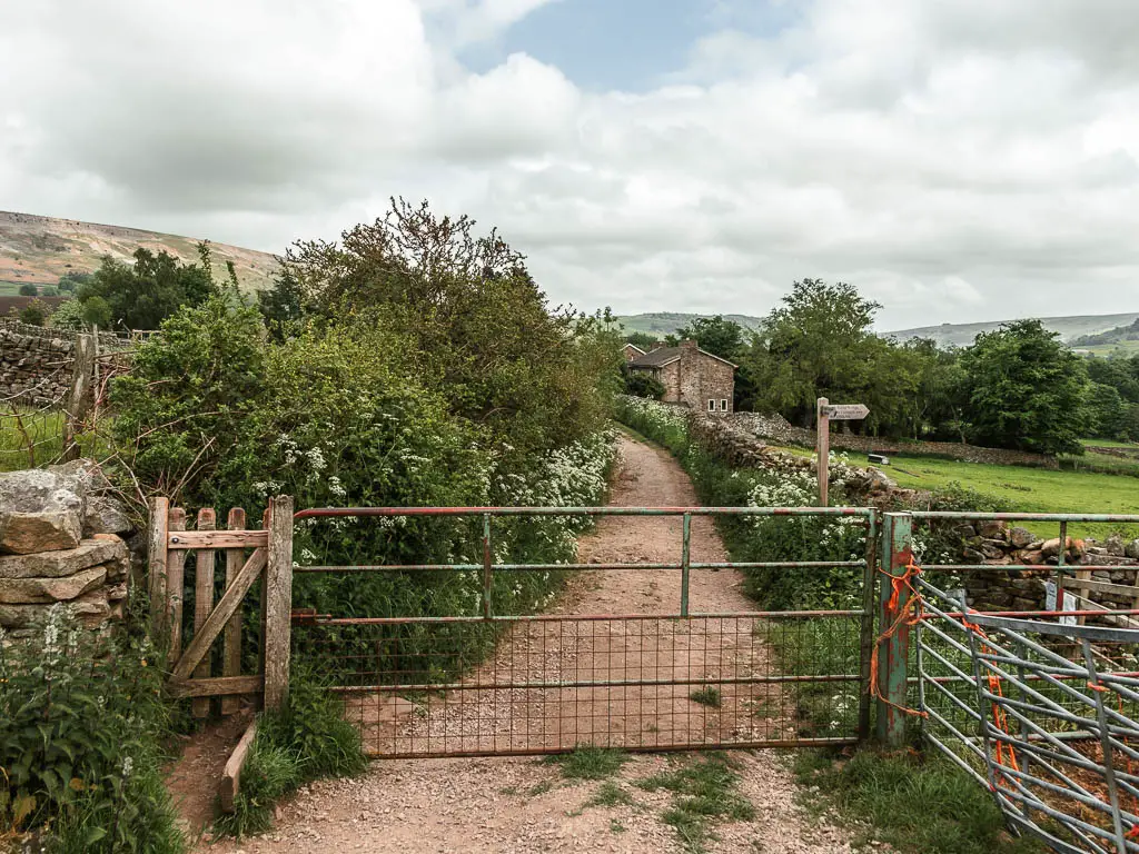 A big metal gate across the path, with bushes lining the left side of the path ahead, and a stone cottage ahead in the distance on the right.