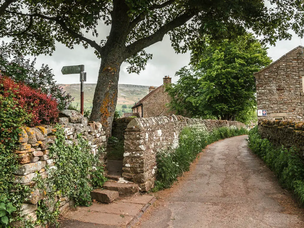 The stone wall lined path leading straight and to the right, with a footpath sign through a gap in the wall to the left, on there walk back into Reeth.