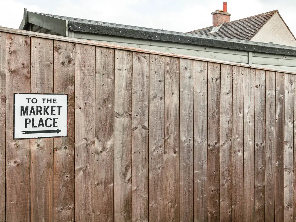 A white sign on the wooden fence, pointing right to walk to the Reeth market place. 