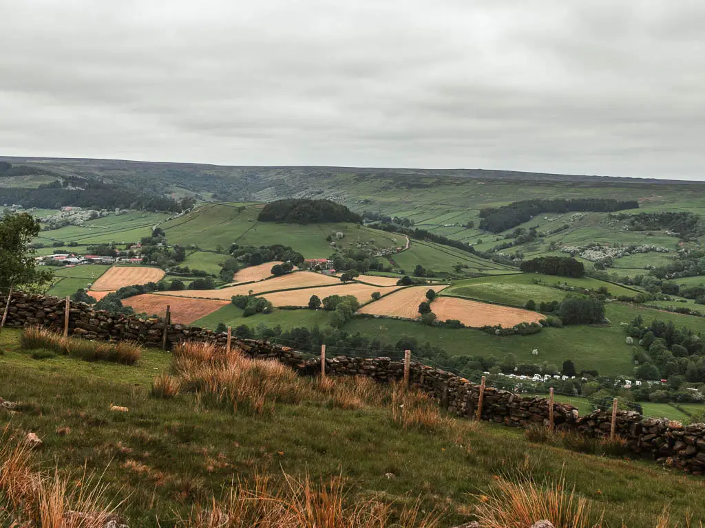 Looking down the hill, to the valley below, on the walk back to Rosedale Abbey. There is a big hill in the middle of the valley. The valley is filled with  green, yellow, and orange patchwork fields.