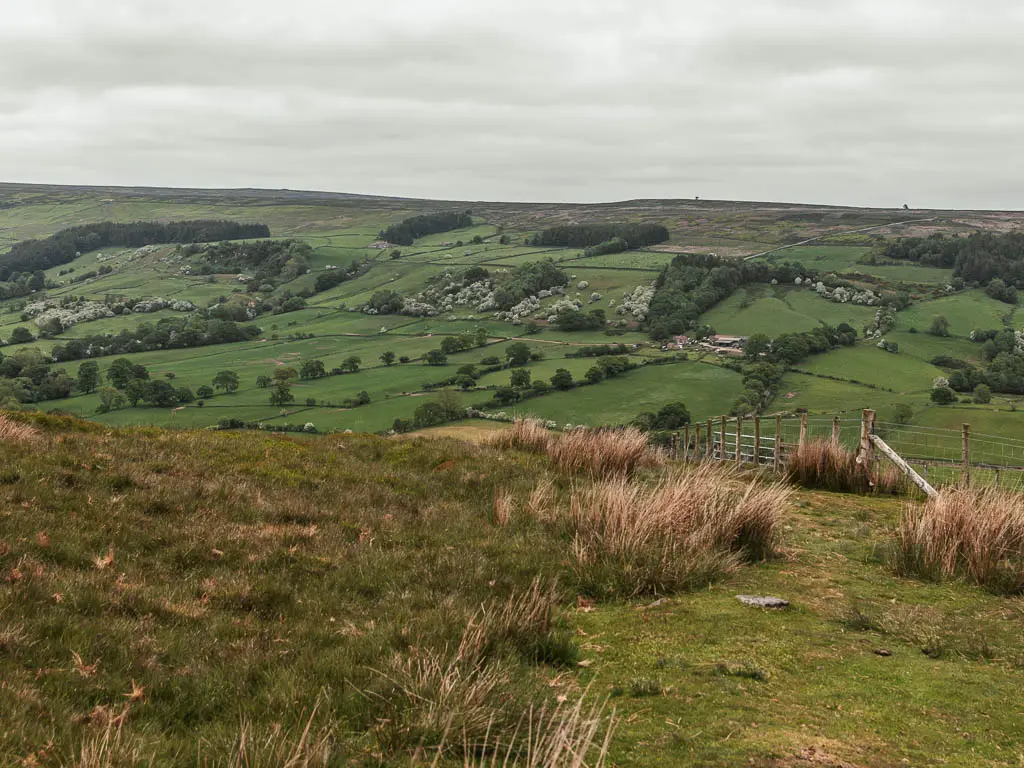 Looking down across the rugged grass field, to the valley below, and hill rising up on the other side.