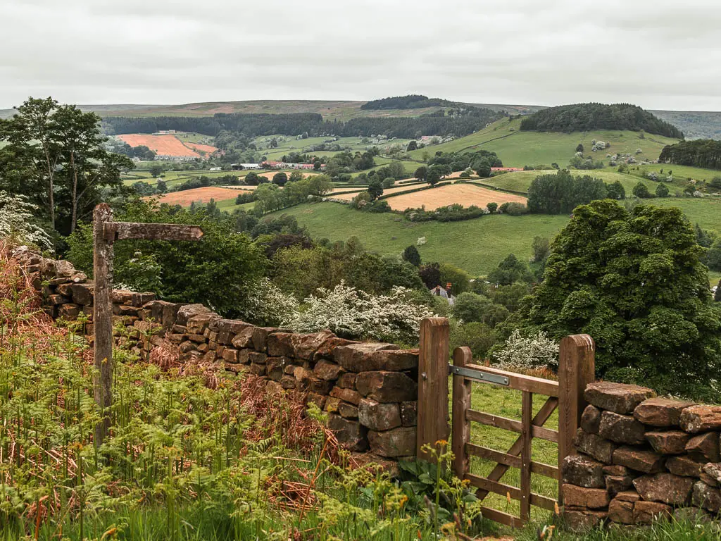 A wooden gate in a stone wall, cutting across there frame, with a view down into the valley below, near the end of the Rosedale Abbey circular walk. There is a wooden trail signpost on this side of the wall, pointing through the gate. The valley is filled with trees, and patchwork fields.