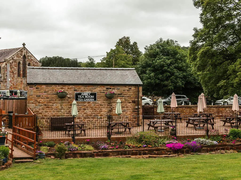 Looking across the neatly cut green to the Rosedale Abbey tea room and garden building at the start of the walk. The budding is made of stone walls. There is a flower patch running across the other side of the green. There are lots of picnic benches next to the tea room building.