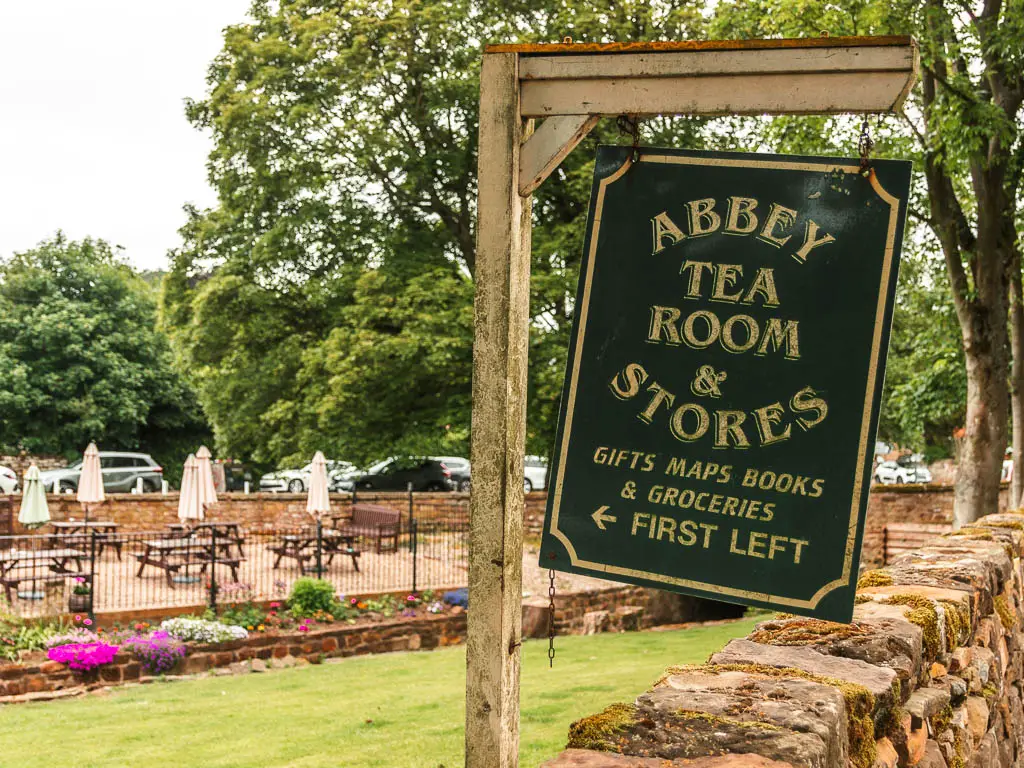 A hanging black sign saying 'Abbey tea room and stores' in gold writing. To the left of the sign is a neatly cut green, with purples flowers lining the other side. Past the flowers is a black railing fence, with picnic benched with closed down white umbrellas.