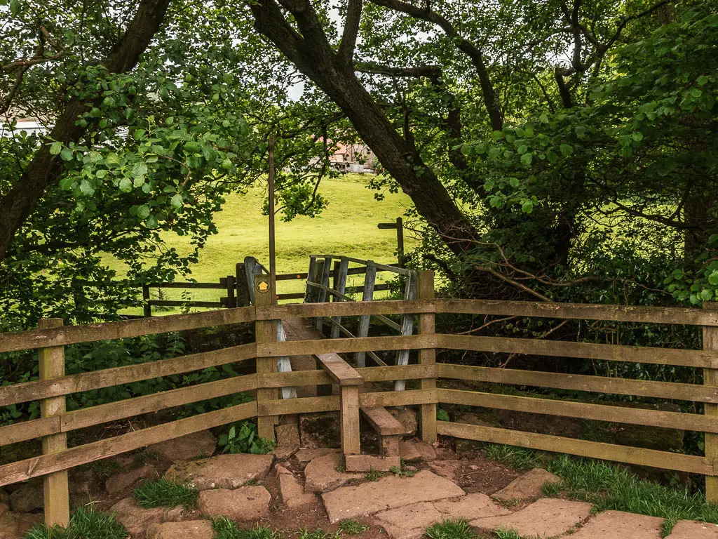 A wooden style in the wooden fence, leading to a bridge. There is a green grass field visible passed the bridge. There are trees surrounding the bridge.