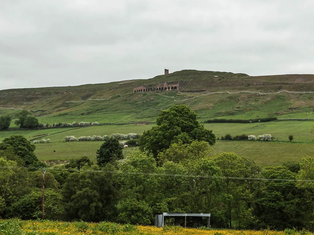 Looking towards a hill, with the ruins of the old kilns and water tower near the top, partway through the walk from Rosedale Abbey. There are lots of green leafy trees at the bottom of the hill.