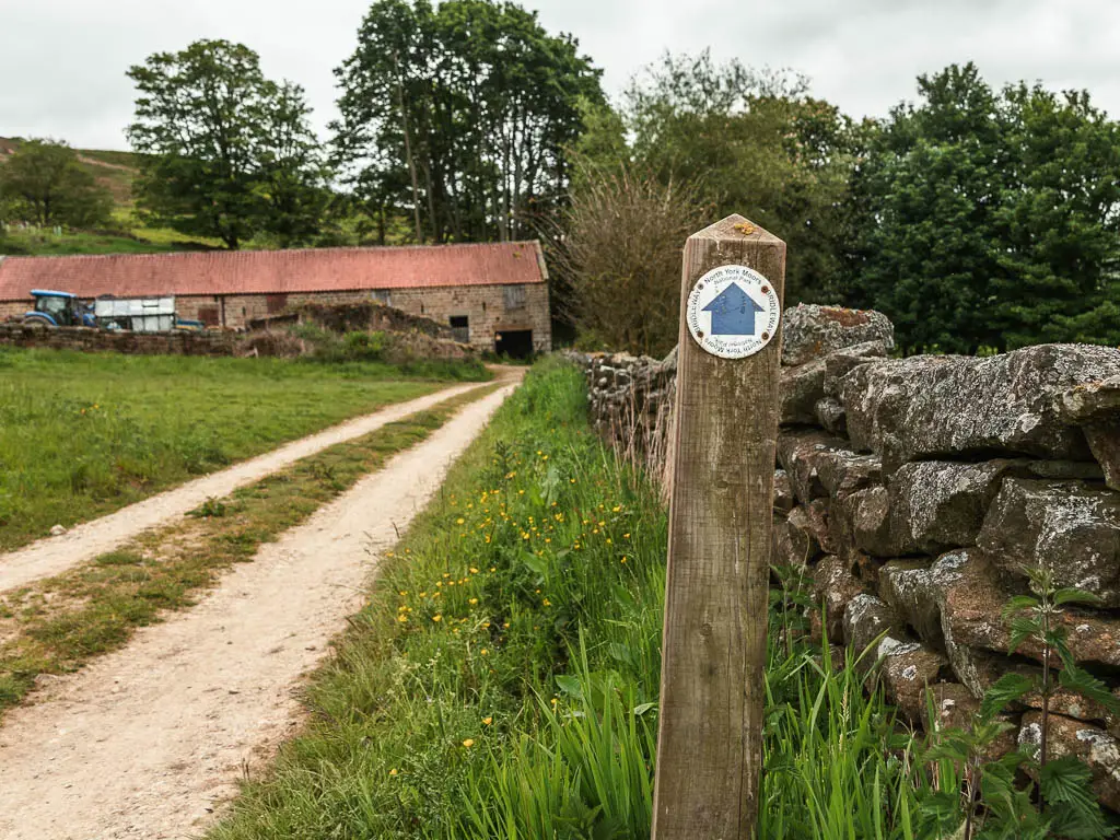 A wooden post with a blue arrow pointing straight ahead. There is a track to the left, and stone wall to the right, and a farm building with a red roof ahead.