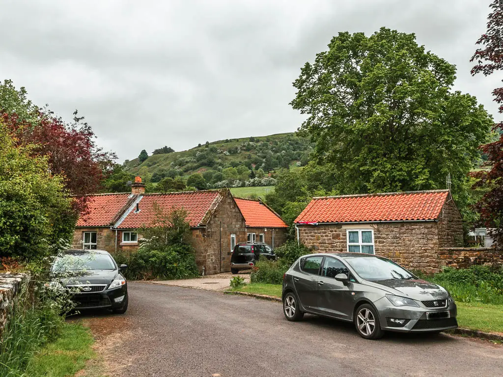 A road, with two parked cars, and some red roofed cottages on the other side of the road. There is a hill in the distance past the rooftops. 