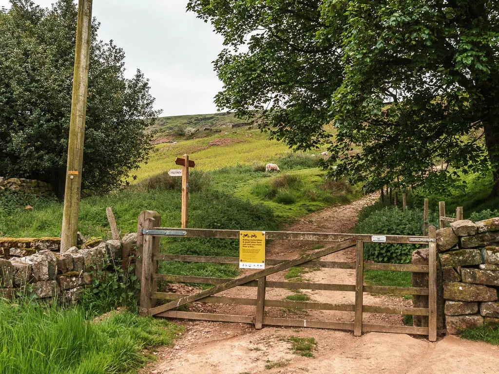 A wooden gate leading to a dirt path up the hill. There is a trail signpost on the left of the path.