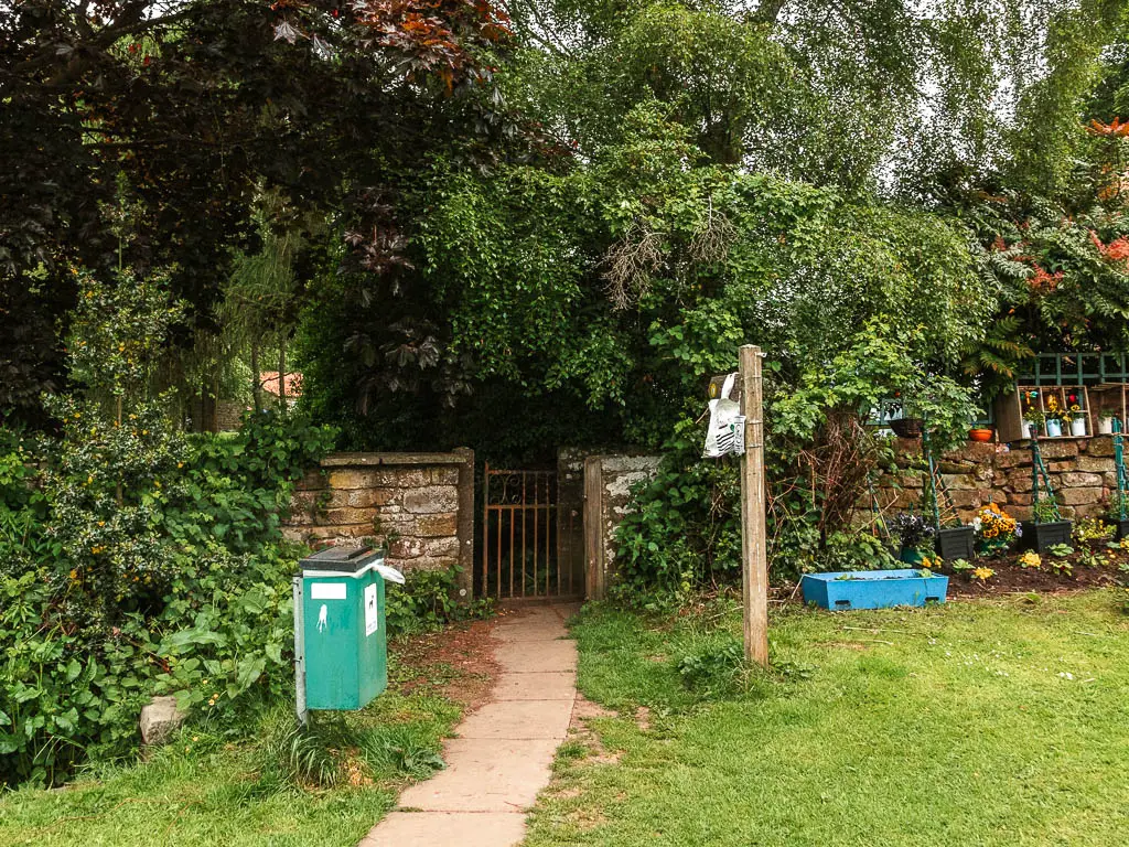 A paved path leading towards a metal gate with trees on the other side. There is a grass area to the right side of the path, and a dog poo pin on the left side.