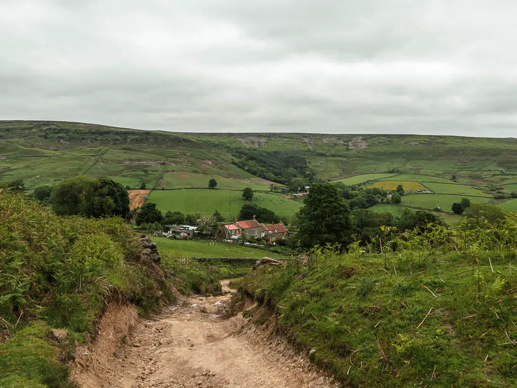 A dirt path leading down hill, lined with grass banks. There is a group of cottages at the bottom of the hill, and a hill rising up beyond them.