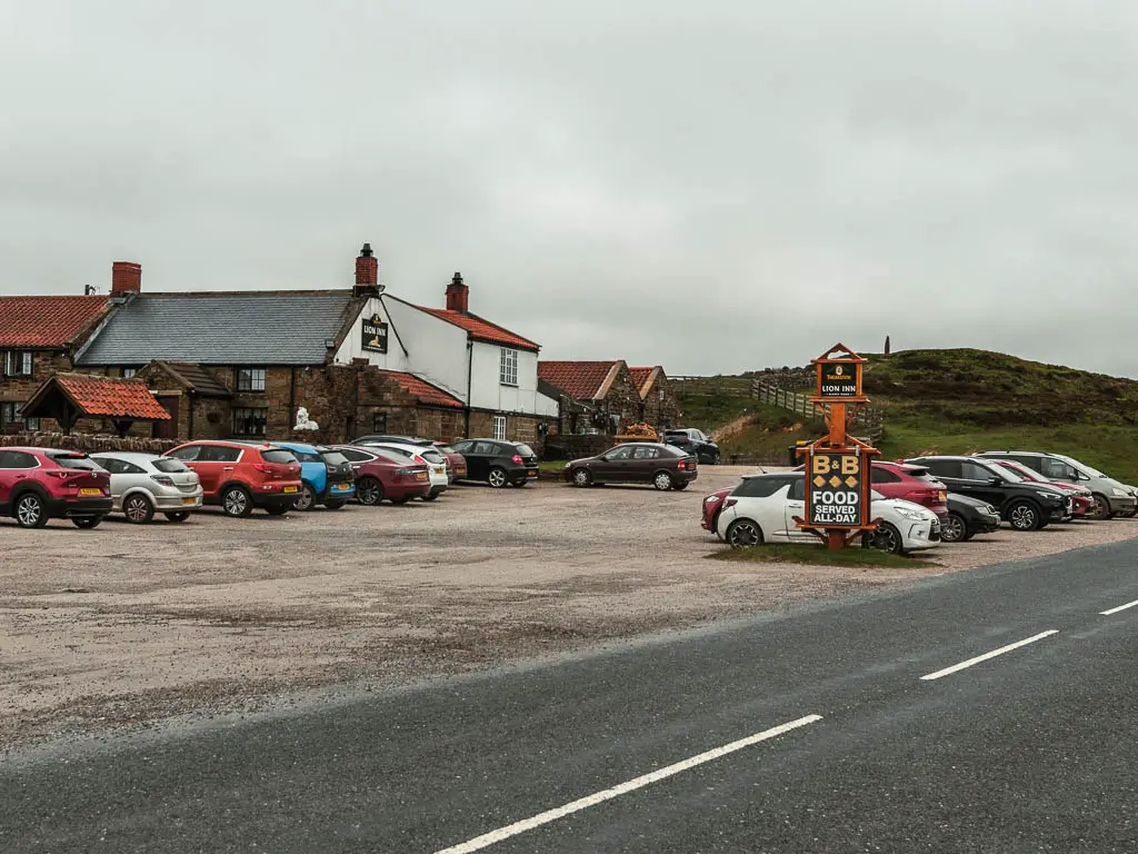 A road, with a large car park filled with parked cars on the other side. There is a pub on the other side of the car park.