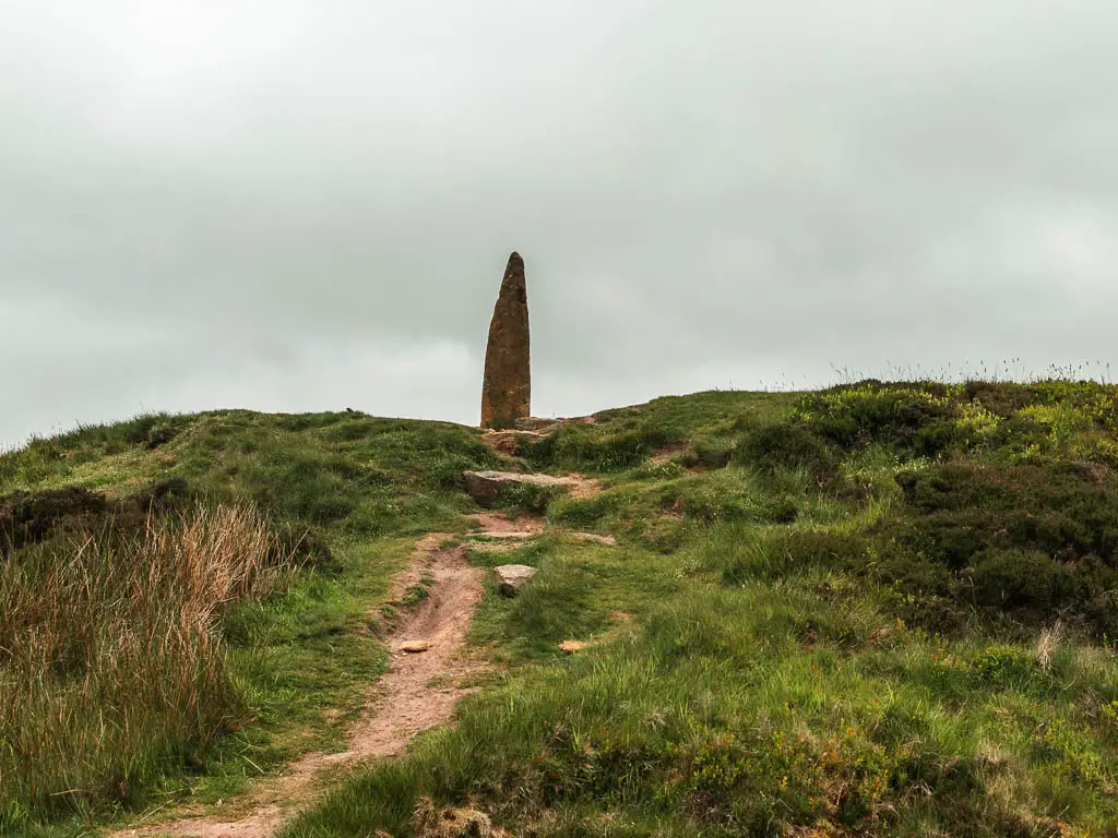 A rugged trail leading up the grass hill to the summit of Blakey Ridge, which has a stone pillar at the top.