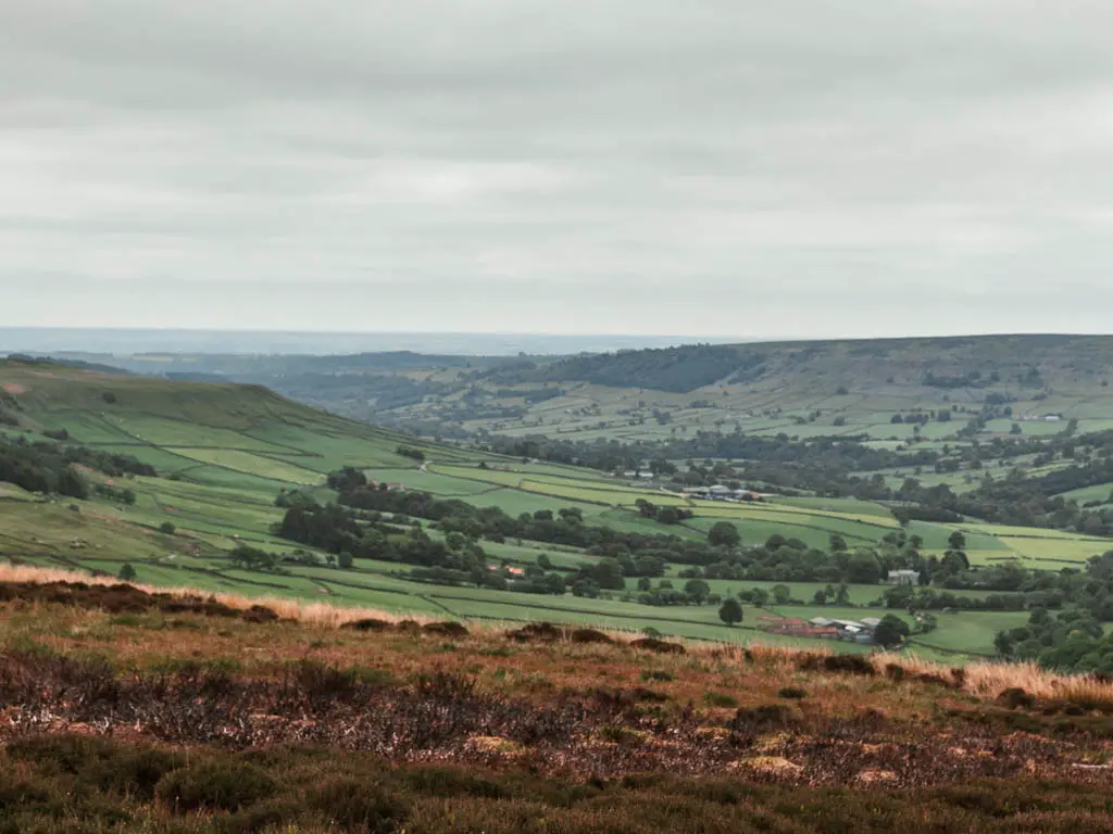 Looking down across the brown moor, to the sweeping valley view ahead. The valley is filled with green grass fields, and trees.