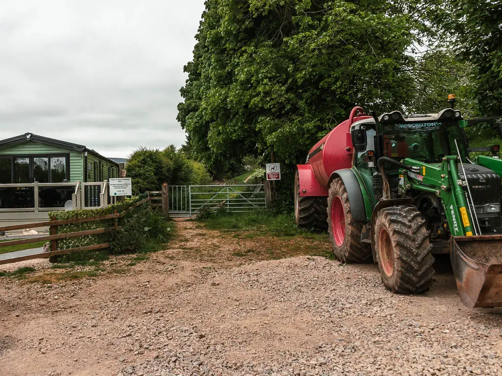 A gravel patch of ground, with a big tractor to the right, and gate ahead, and a caravan trailer to the left.