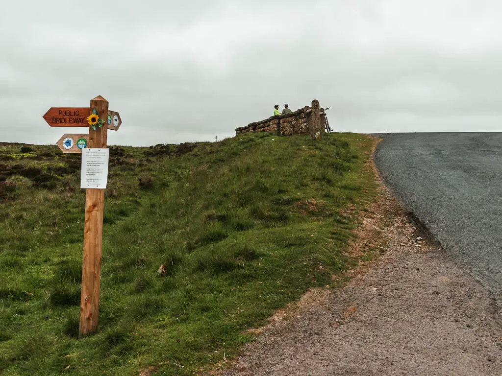 A road leading uphill to the right, with a grassy area to the left. There is a wooden footpath sign on the green, pointing to the left, and ahead.