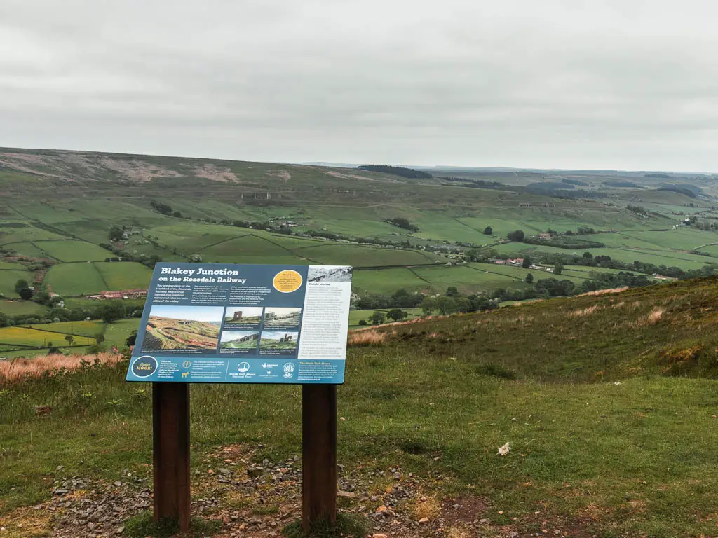 An information board marking Blakey Junction and the Rosedale Railway, on the walk back towards Rosedale Abbey. The information board is on a green area, with a view to the valley hills in the distance.