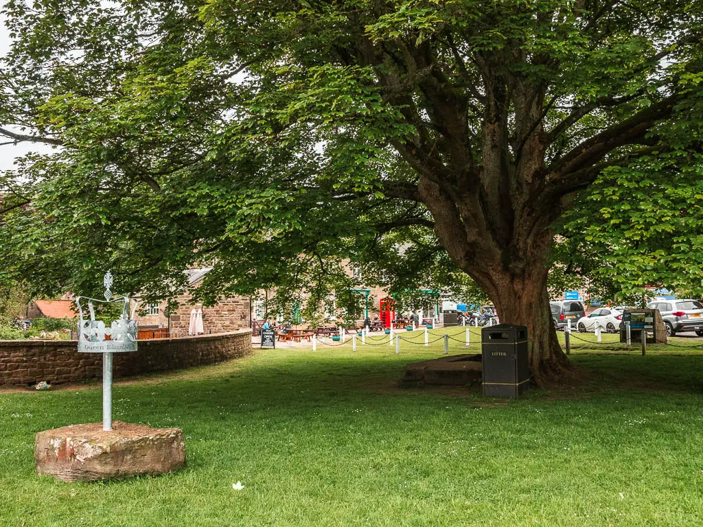 A green area, with a big tree in the middle. There is a crown statue to the left.